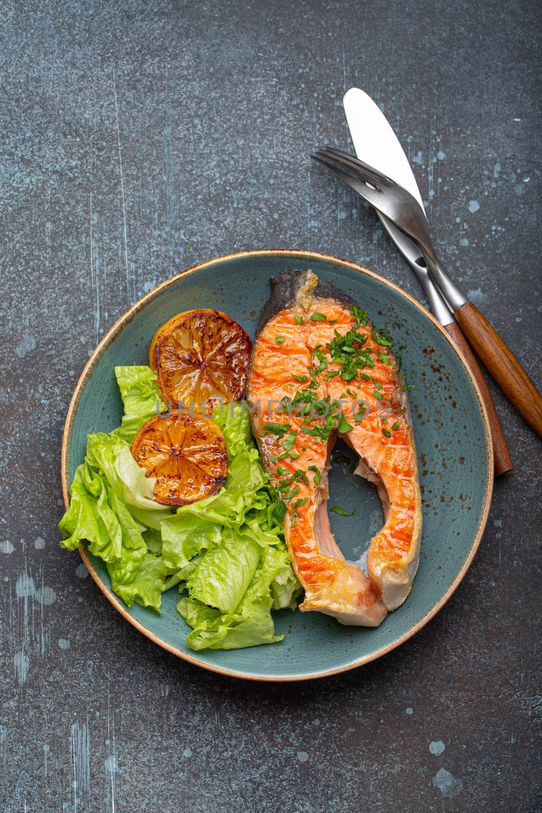 Grilled fish salmon steak and green salad with lemon on ceramic plate on rustic blue stone background top view, balanced diet or healthy nutrition meal with salmon and veggies.
