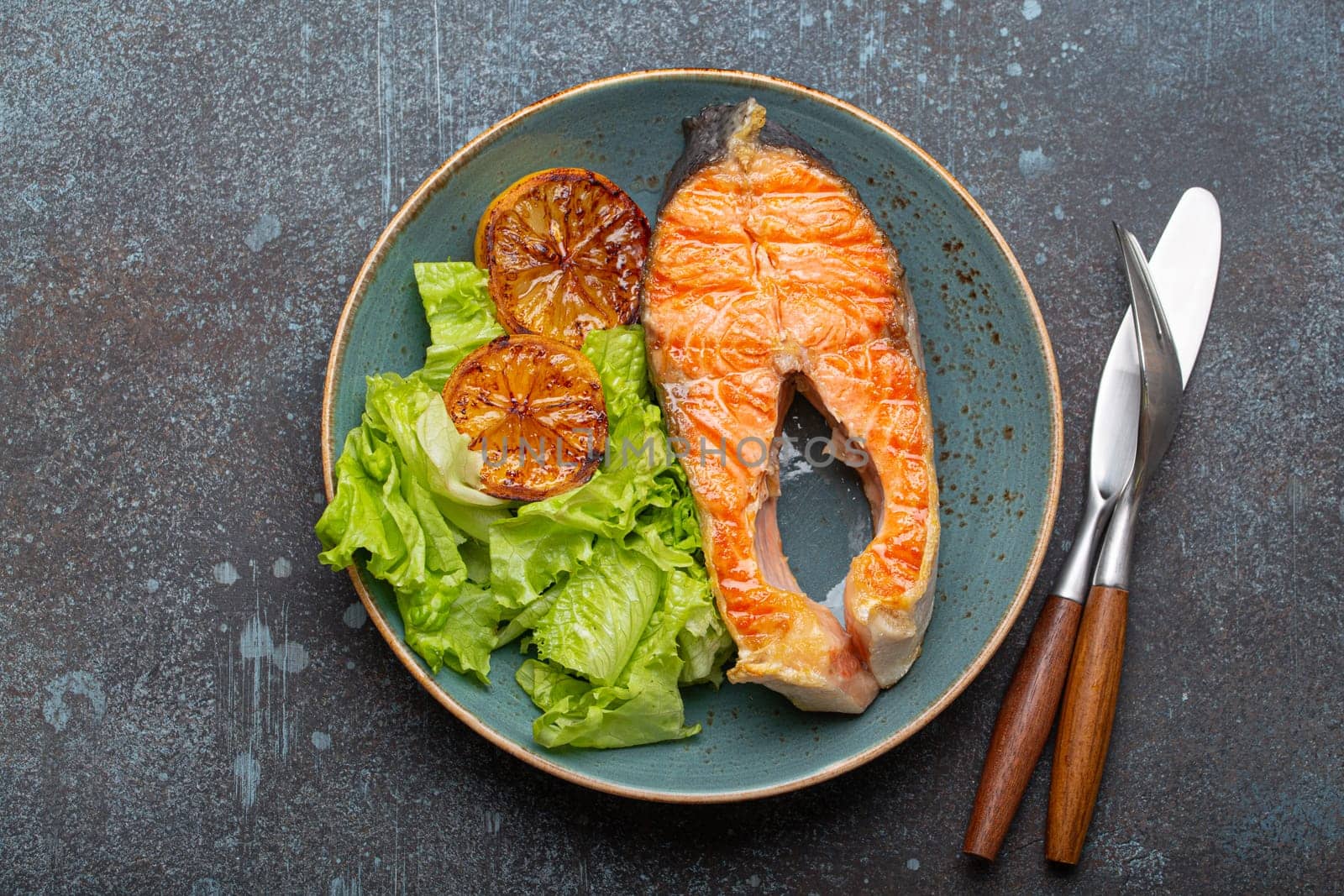 Grilled fish salmon steak and green salad with lemon on ceramic plate on rustic blue stone background top view, balanced diet or healthy nutrition meal with salmon and veggies by its_al_dente