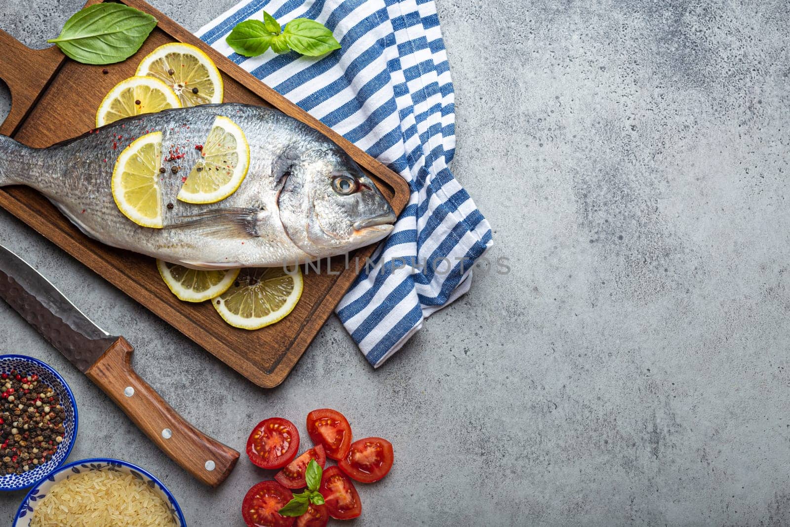 Raw fish dorado with ingredients lemon, fresh basil, cut cherry tomatoes, uncooked rice on wooden cutting board with knife on rustic stone background top view, cooking healthy fish dorado. Copy space