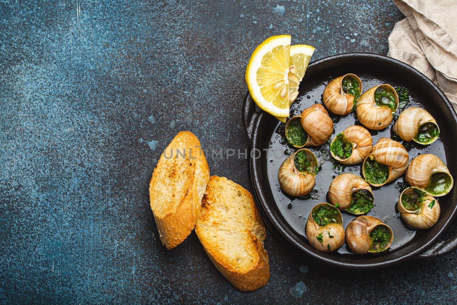 Escargots de Bourgogne Snails with Garlic Butter and Parsley in black cast iron pan with Lemon and Toasted Baguette Slices on rustic stone background top view, traditional French Delicacy, copy space. by its_al_dente