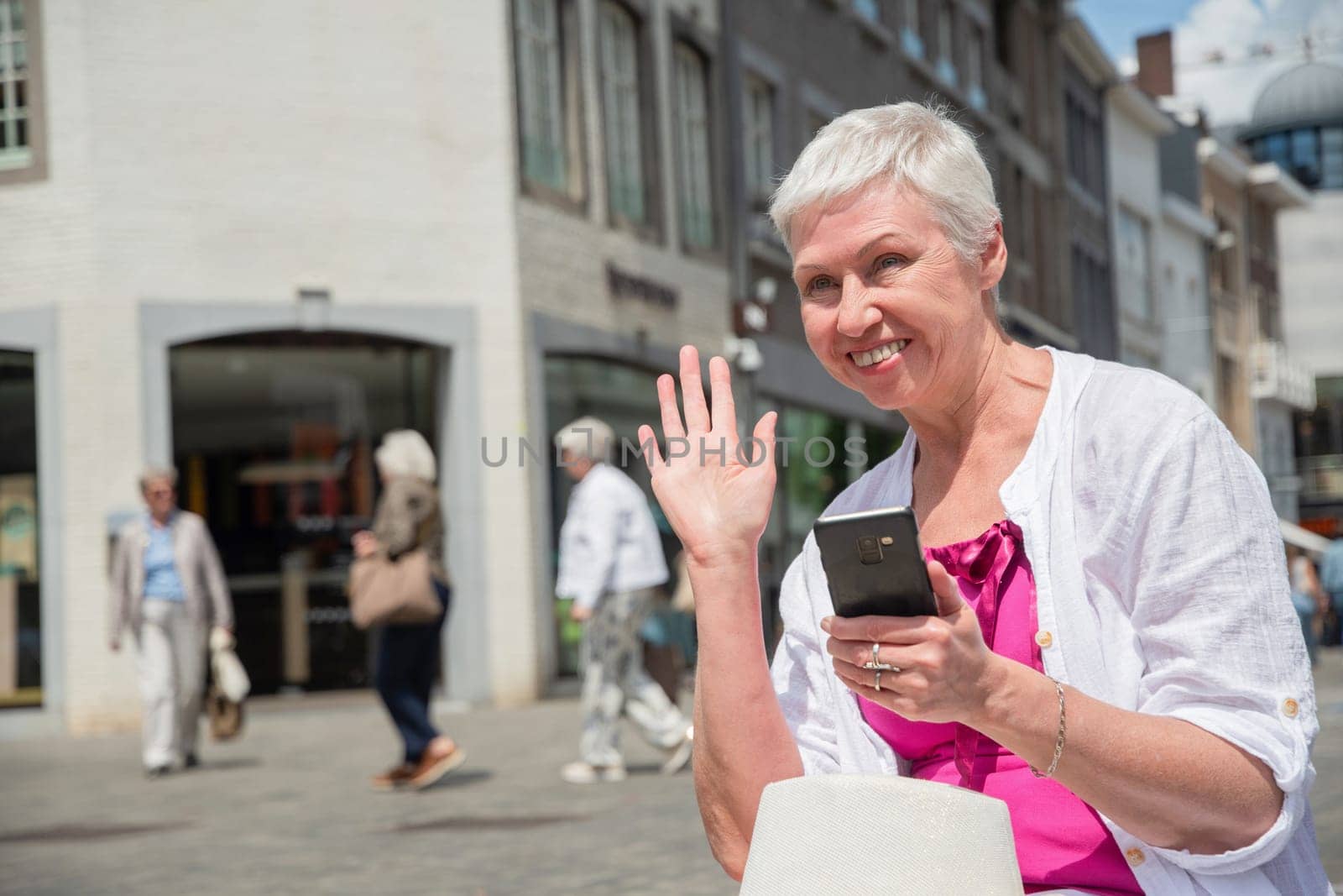 a sixty year old caucasian woman in a hat and stylish summer clothes waves to a passerby and smiles and holds a smartphone in his other hand, sits on a bench in the city, High quality photo