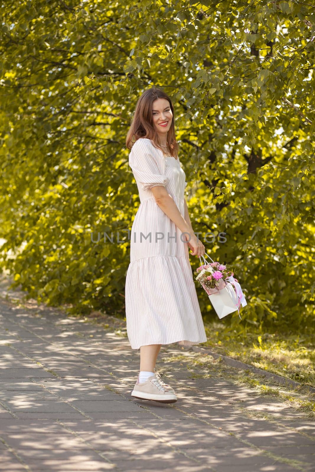 Beautiful girl with flowers, in a dress, glasses and sneakers. A beautiful bouquet of flowers in a box in the hands of a beautiful girl who walks along the street on a sunny day.