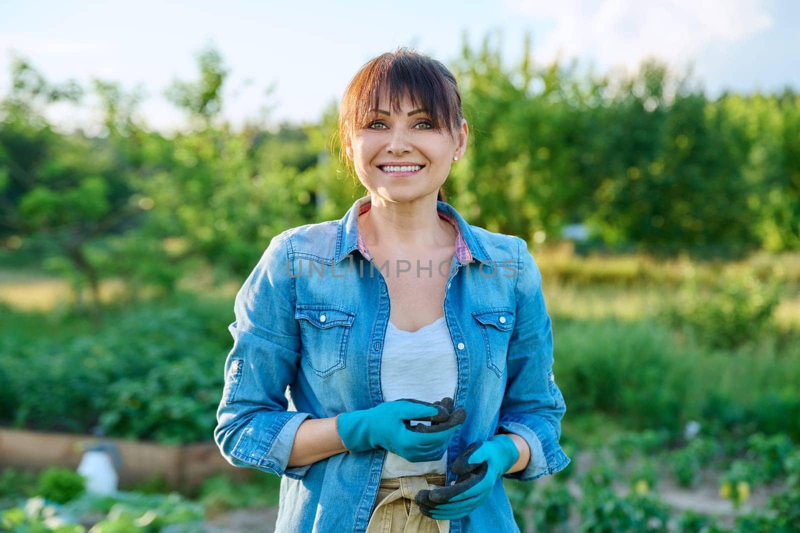 Portrait of smiling woman gardener farmer, nature field with plants vegetables background. Middle-aged female in gloves looking at the camera. Farming, agriculture, small business concept