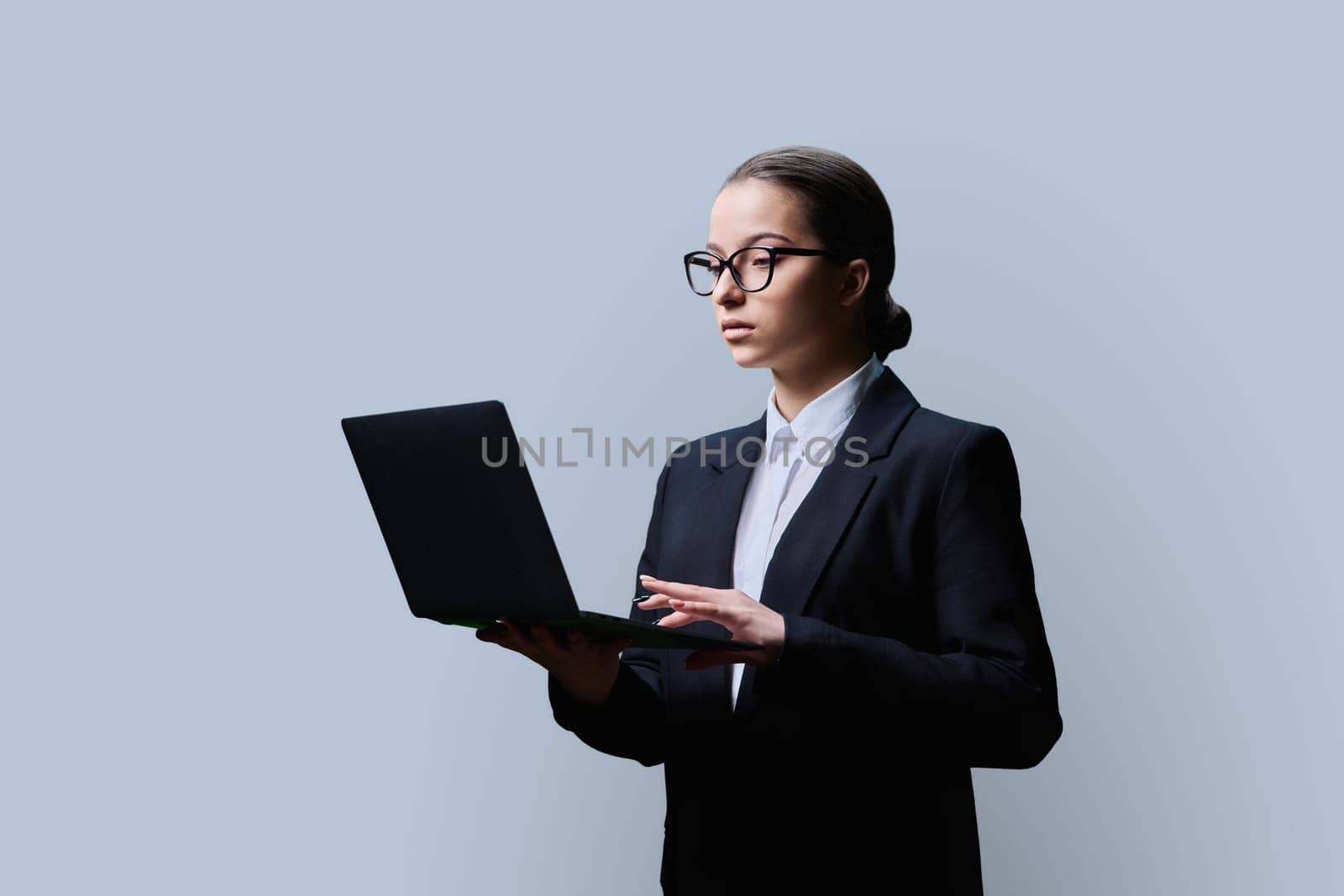Serious teenage female student in formal business style using computer laptop, on grey studio background. Digital technologies, education, virtual educational services, online learning, youth concept