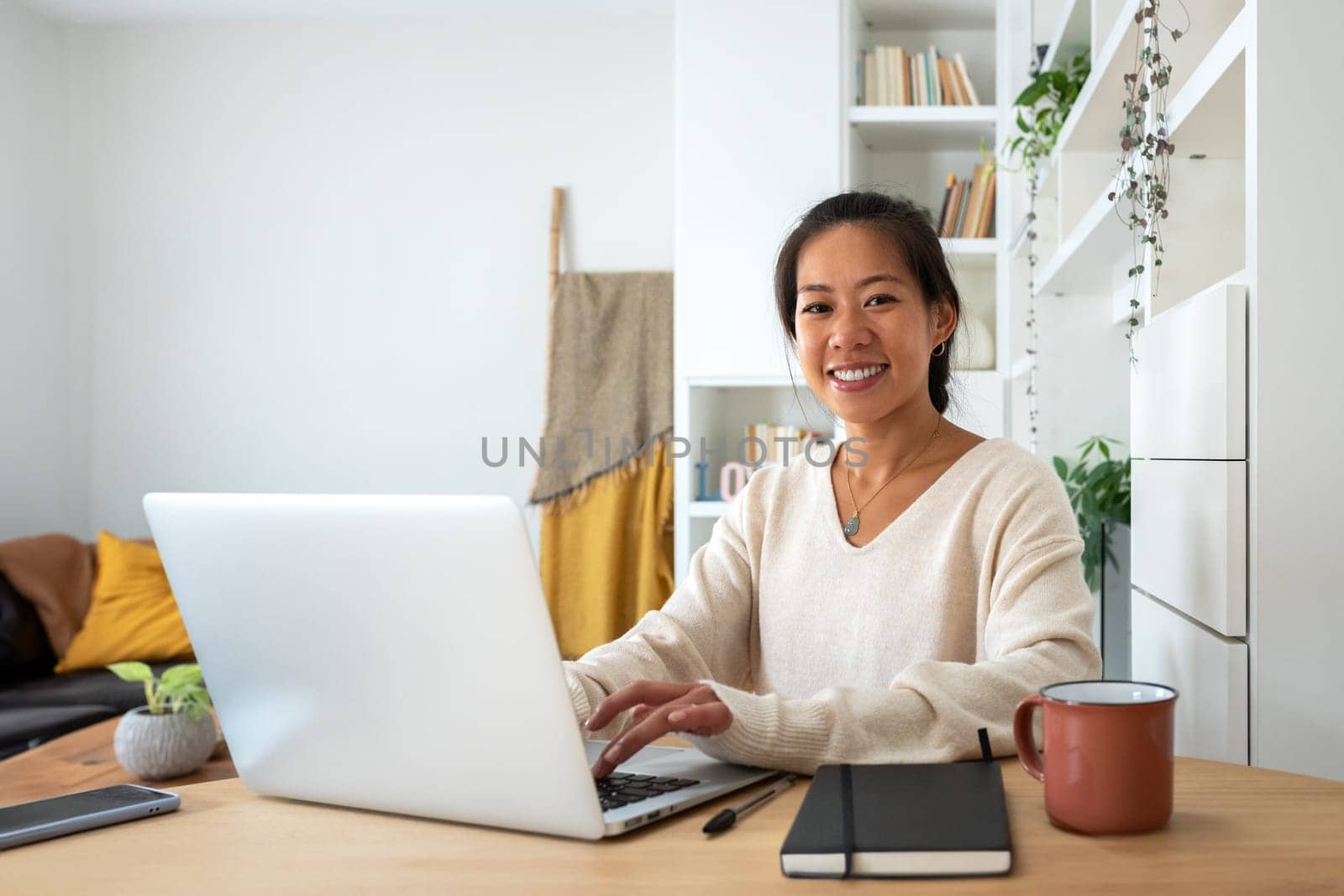 Happy Asian female entrepreneur working using laptop looking at camera. Smiling Chinese woman working with computer at home office. Technology concept.