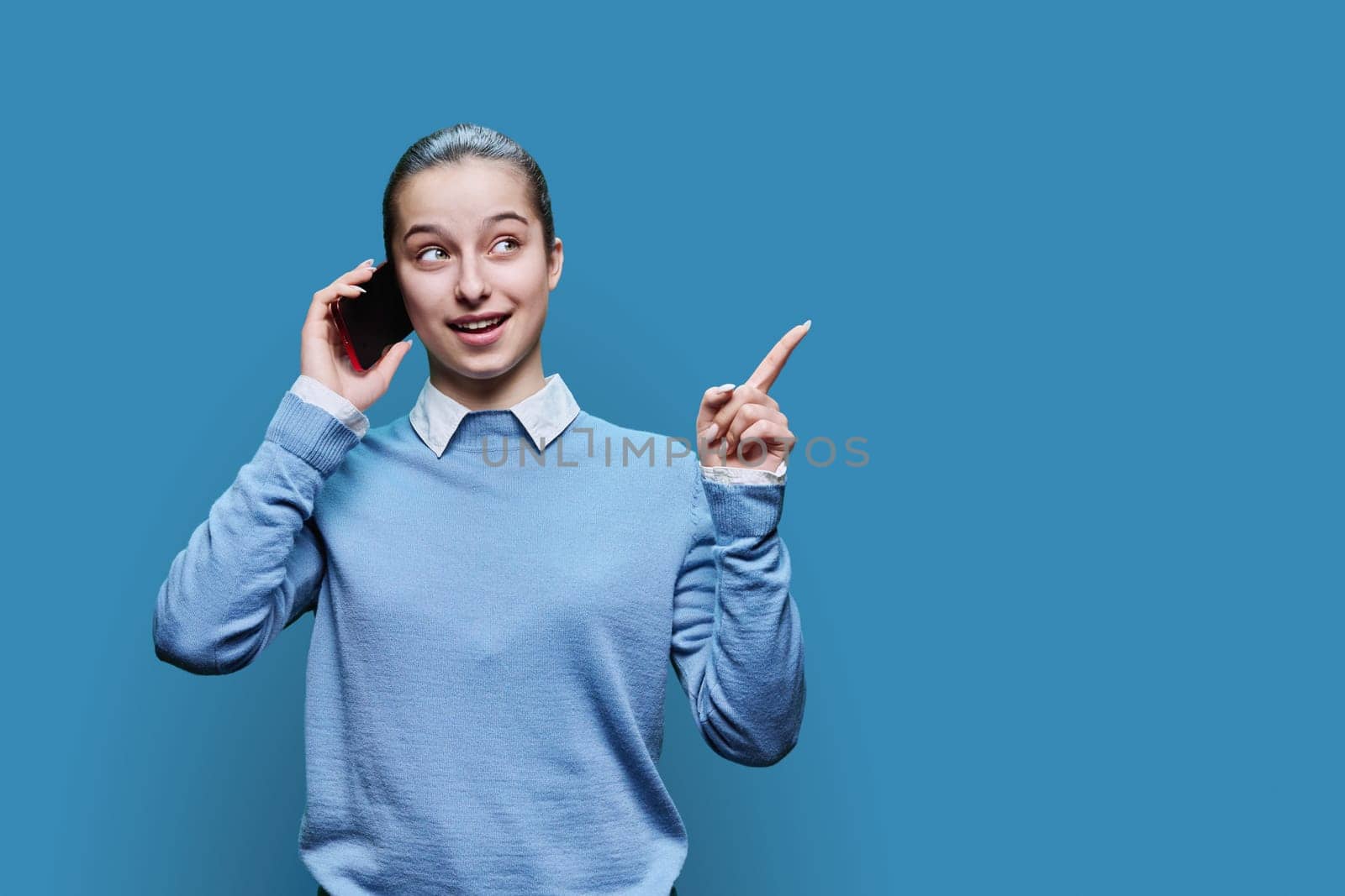 Joyful emotional surprised happy cheerful teenage student, girl 15, 16 years old talking on mobile phone on blue studio background. Lifestyle, communication, adolescence concept