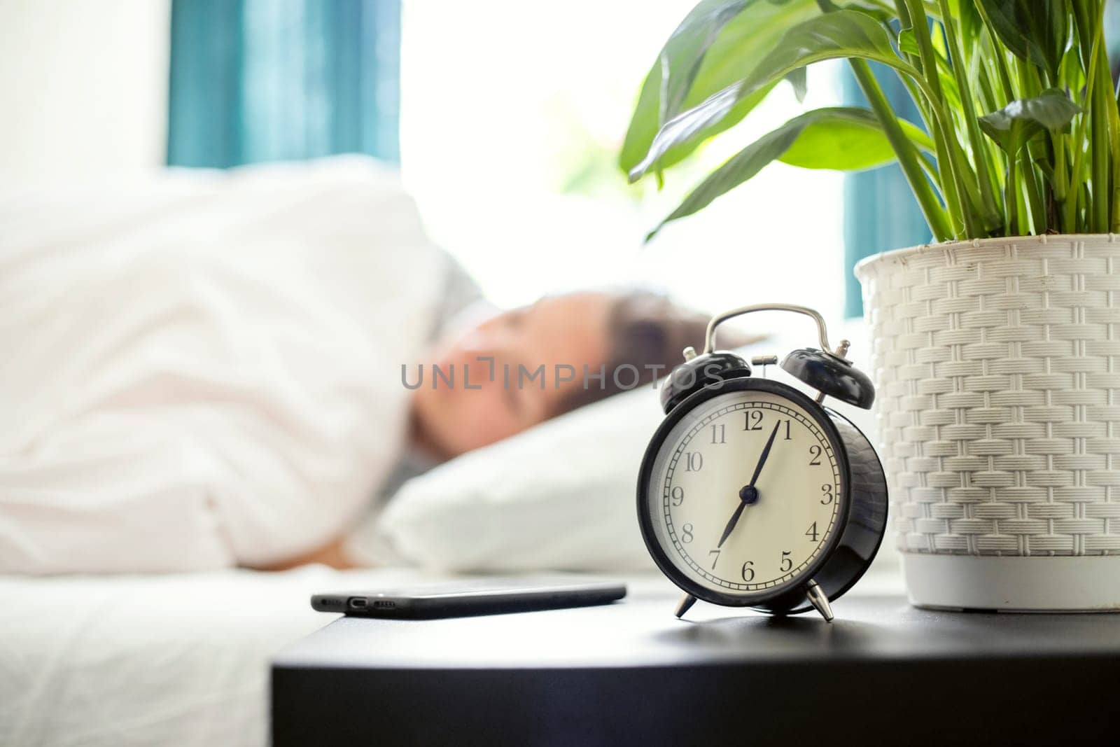 Alarm clock standing on bedside table going to ring early morning to wake up woman in bed sleeping in background