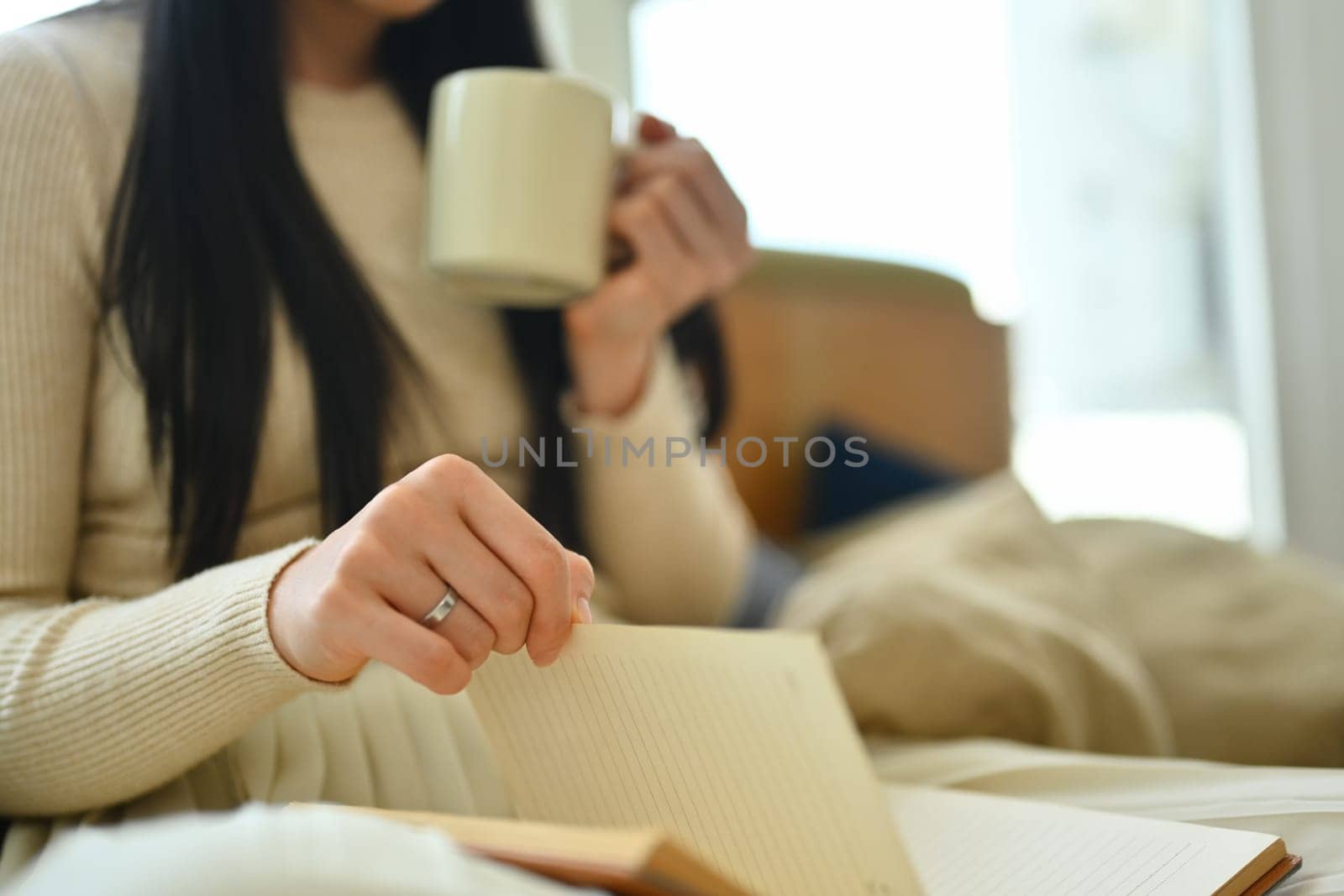 Calm young woman in warm sweater drinking hot tea and reading book in living room.