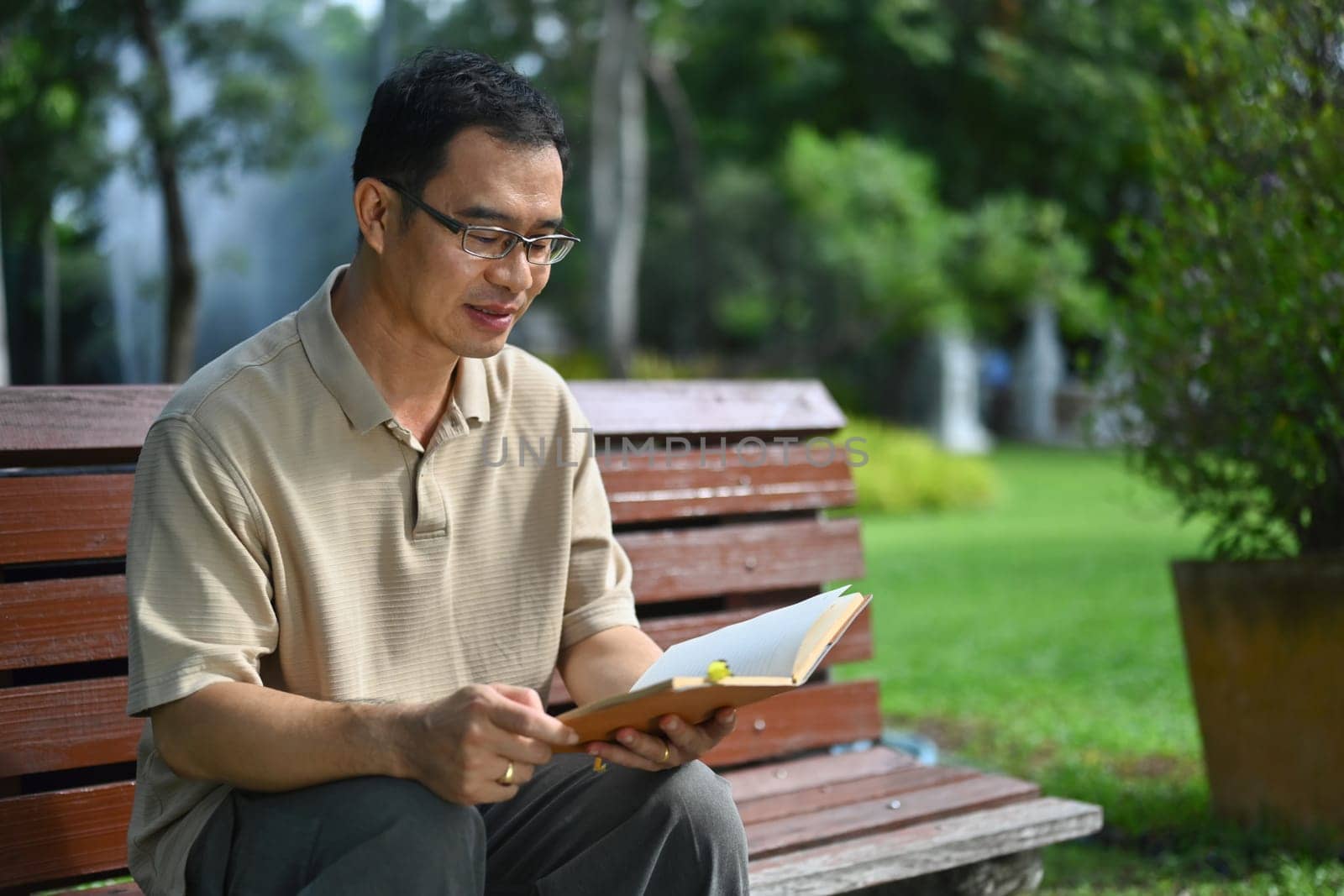 Pleased middle age man reading favorite novel while sitting on bench at public park by prathanchorruangsak