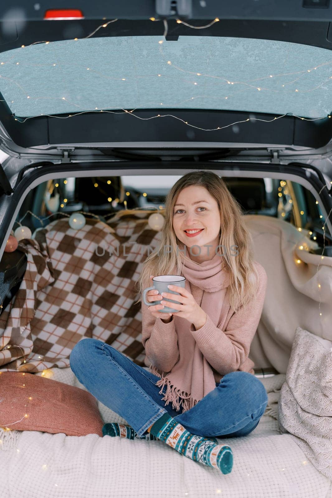 Smiling girl with a cup of coffee sitting cross-legged in the trunk of a car on a blanket by Nadtochiy