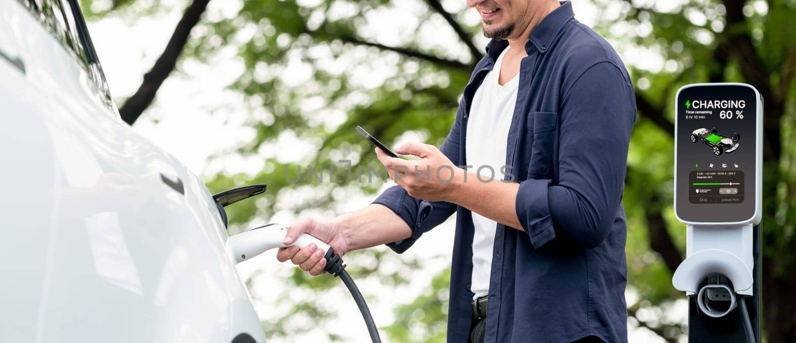 Man using smartphone online banking application to pay for electric car battery charging from EV charging station during vacation road trip at national park or summer forest. Panorama Exalt