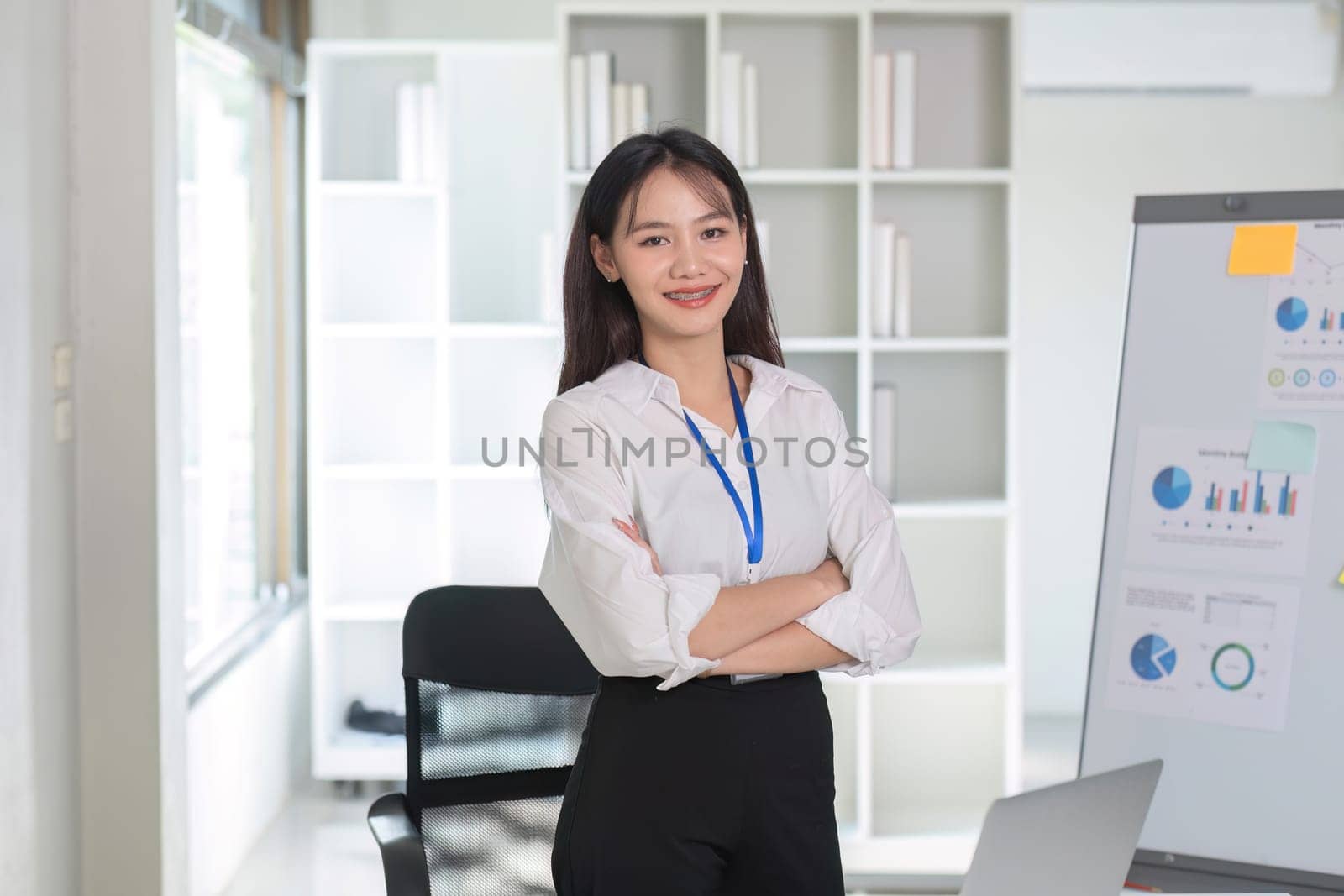 Portrait of an attractive Asian businesswoman standing with her arms crossed in front of a work plan showing financial information, graphs and taxes..