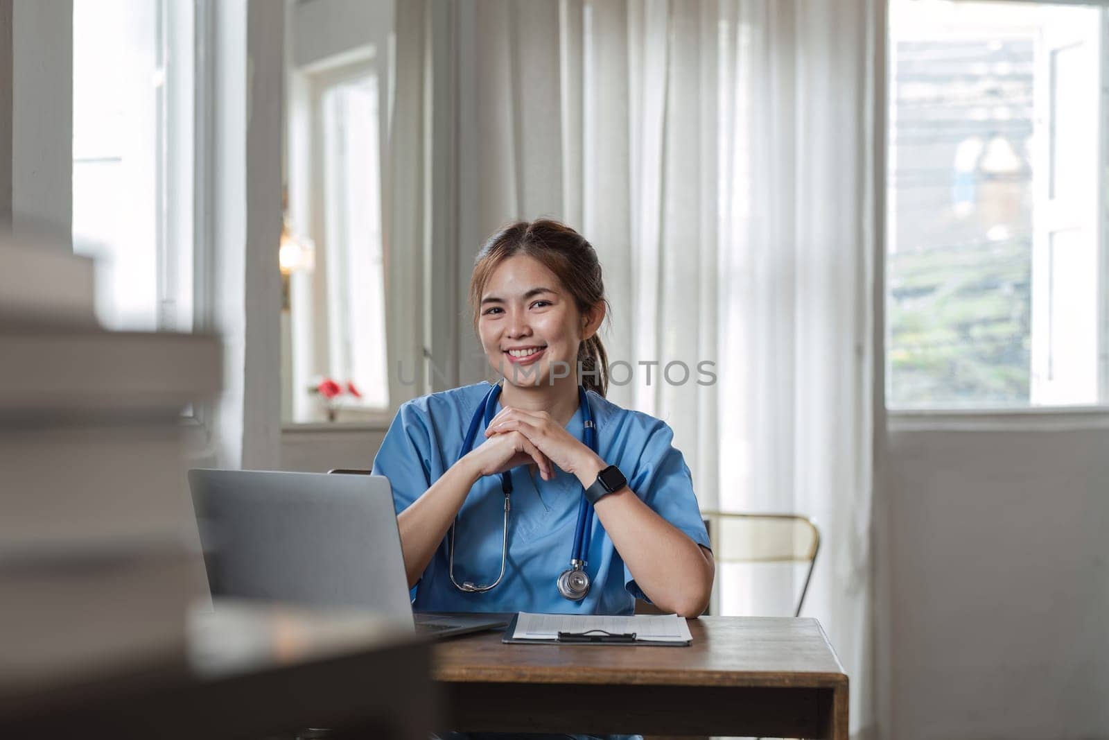 young asian lady doctor in white medical uniform with stethoscope using computer laptop talking video conference call with patient at desk in health clinic or hospital. by wichayada