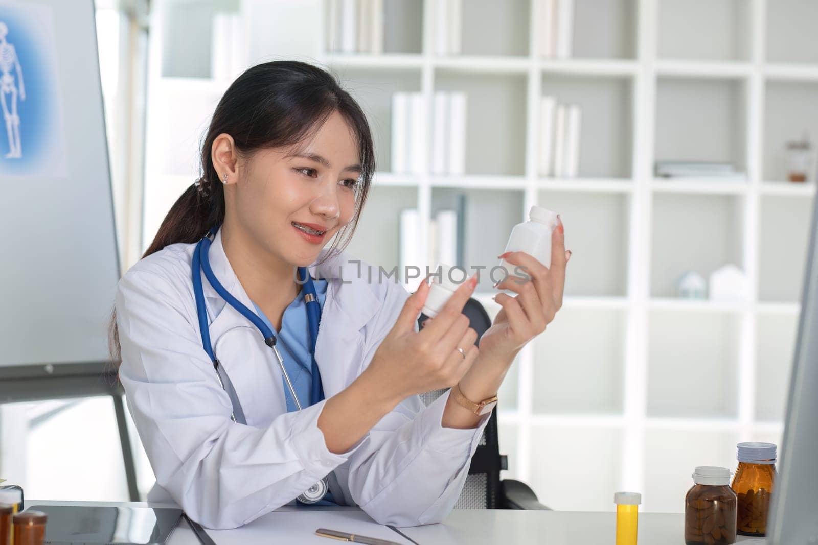 Asian female doctor holding pill bottle in hand, writing prescription, sitting at prescription table, pharmaceutical concept. by wichayada