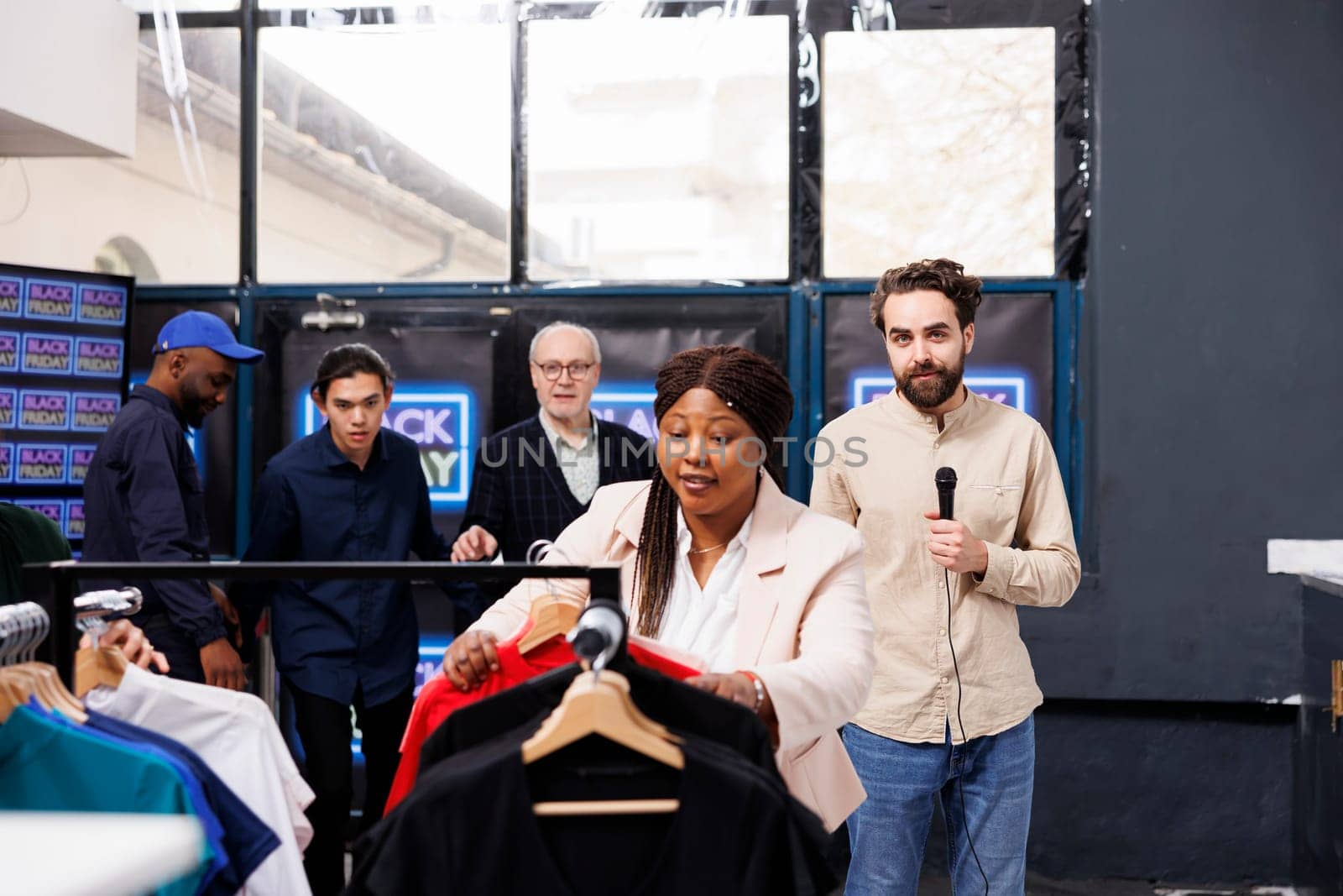 Journalist holding mic looking at camera while reporting on Black Friday sales from shopping mall. Crowd of excited people shoppers running past TV reporter into fashion outlet to get best deals