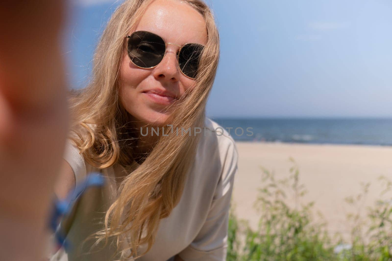 Young woman in sunglasses with shell bracelets looking into camera. Influencer Using mobile phone for video call selfie waving on the beach seacoast. Holiday vacation advertisement concept. Social media