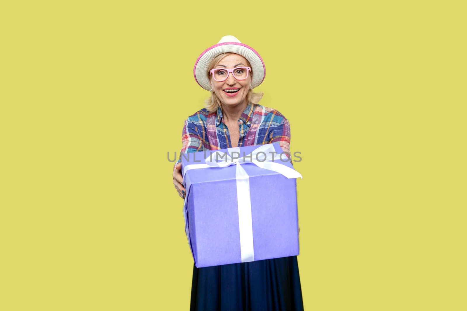 Portrait of excited positive mature woman wearing checkered shirt, hat and eyeglasses giving present box, congratulating with holiday. Indoor studio shot isolated on yellow background.