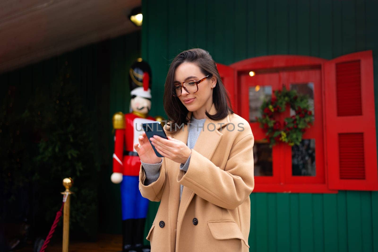 Smiling business woman wearing trench coat standing on Christmas city street using smartphone applications on cell phone, reading news on smartphone, fast connection, checking mobile apps outdoors.