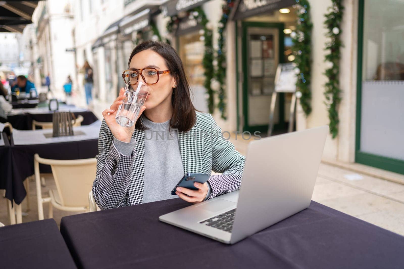Freelancer using smartphone and laptop sitting outdoor cafe drink water wearing glasses, working in cafe on laptop, holding mobile phone in hands, drinking water from glass.