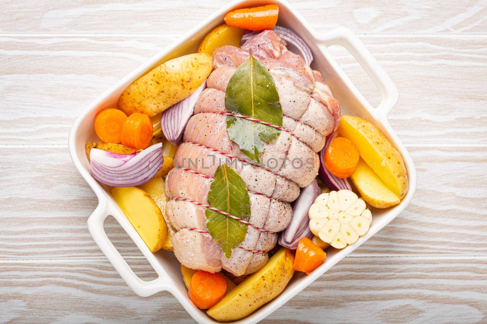 Raw wrapped rolled sliced pork in white casserole dish with potatoes, vegetables and herbs on rustic white wooden background top view. Pork roll with vegetables ready to be prepared by its_al_dente