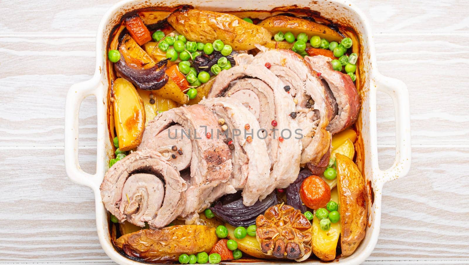 Rolled sliced pork roasted in white casserole dish with potatoes, vegetables and herbs on rustic white wooden background top view. Baked pork roll with vegetables for dinner by its_al_dente