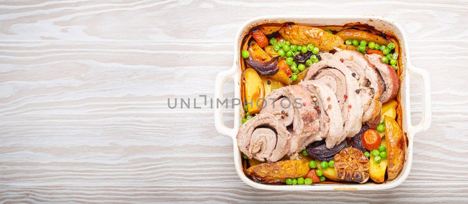 Rolled sliced pork roasted in white casserole dish with potatoes, vegetables and herbs on rustic white wooden background top view. Baked pork roll with vegetables for dinner by its_al_dente