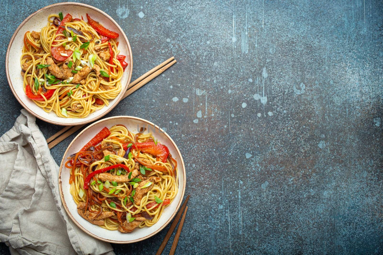 Two bowls with Chow Mein or Lo Mein, traditional Chinese stir fry noodles with meat and vegetables, served with chopsticks top view on rustic blue concrete background, space for text.