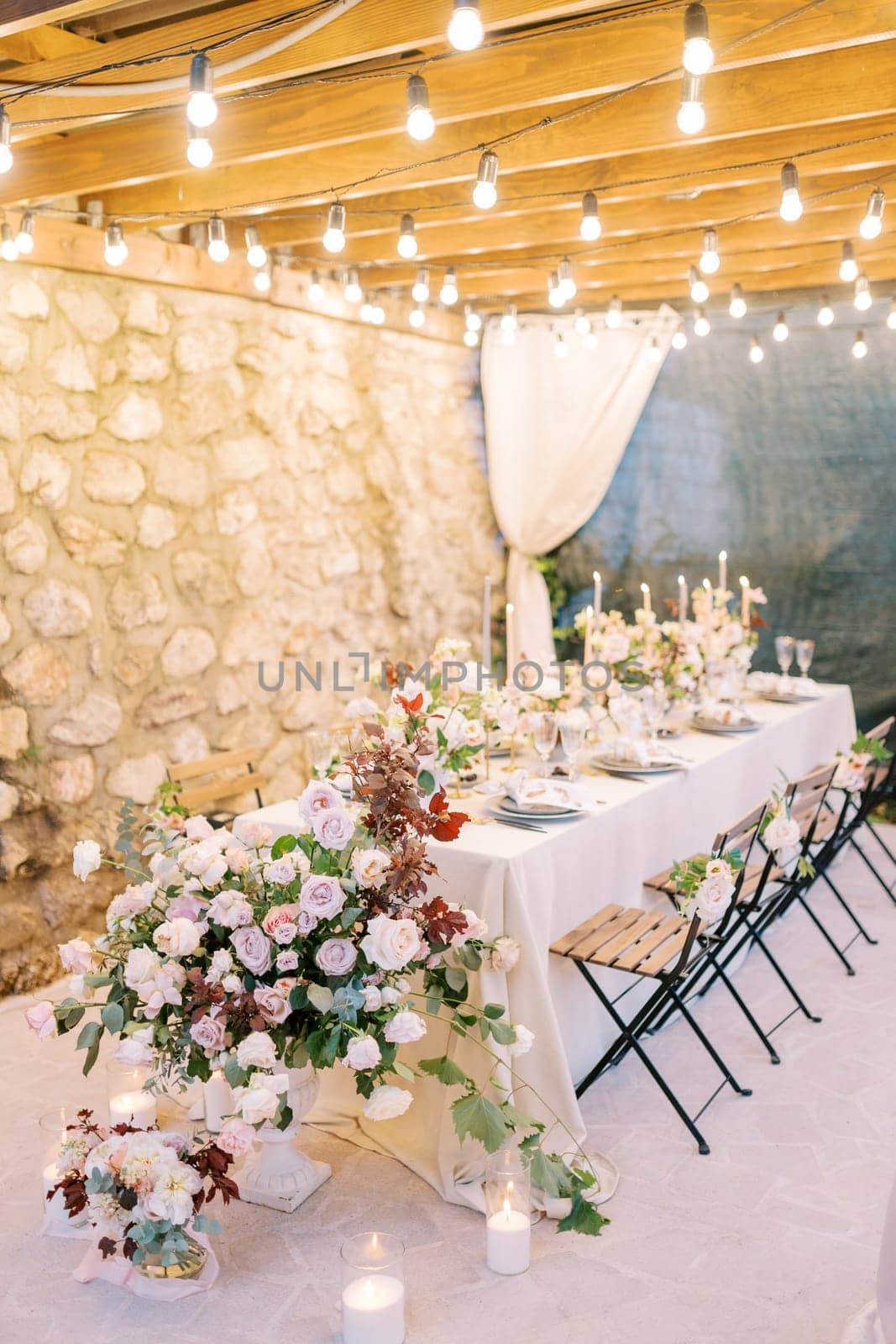 Bouquets of flowers and lit candles stand on a festive table on the terrace in the light of garlands by Nadtochiy