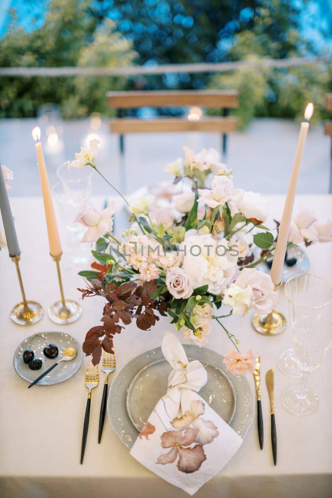 Lighted candles stand on the table near a bouquet of flowers in front of a plate with a knotted napkin by Nadtochiy