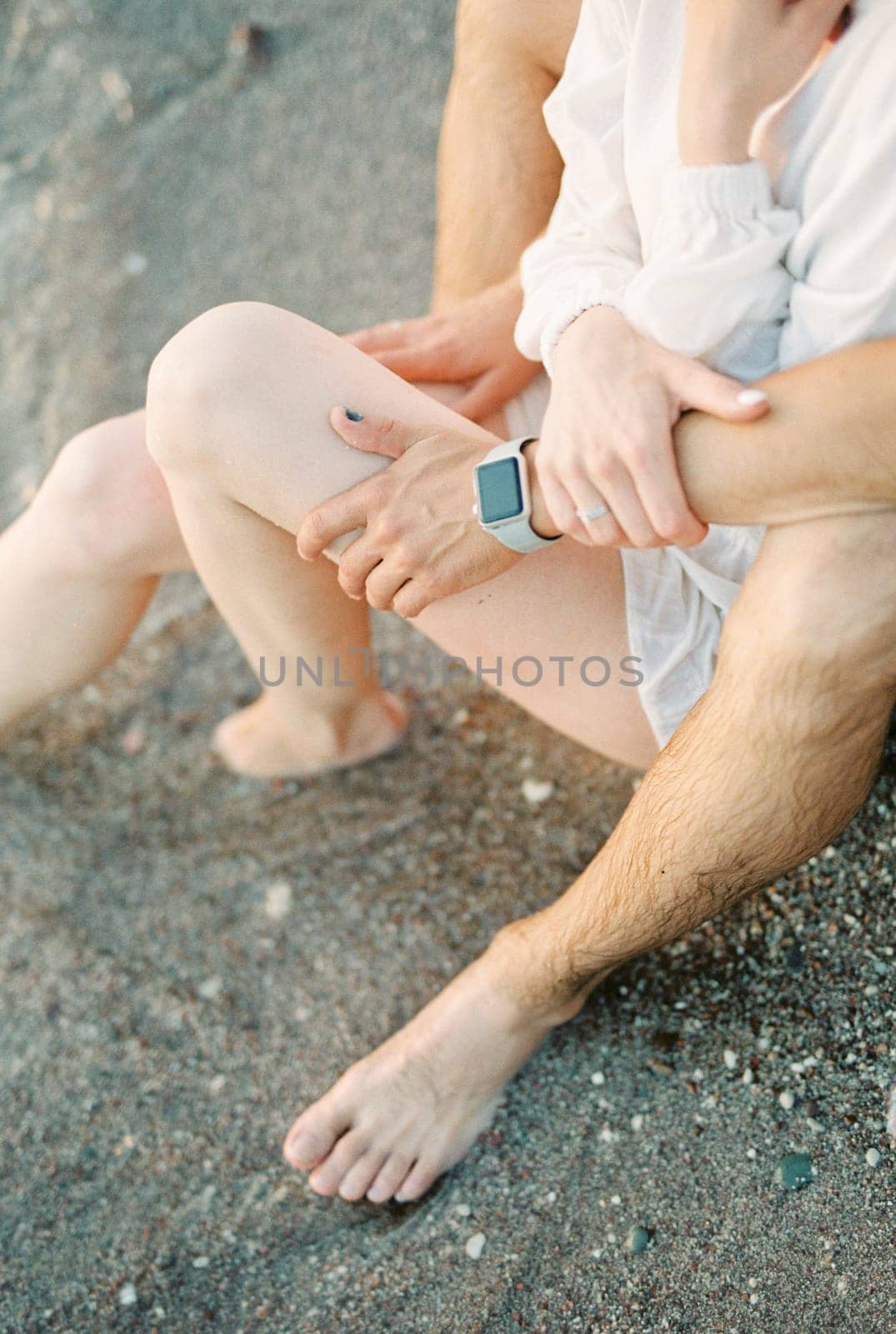 Man hugs woman legs while sitting on the seashore. Cropped. Faceless by Nadtochiy