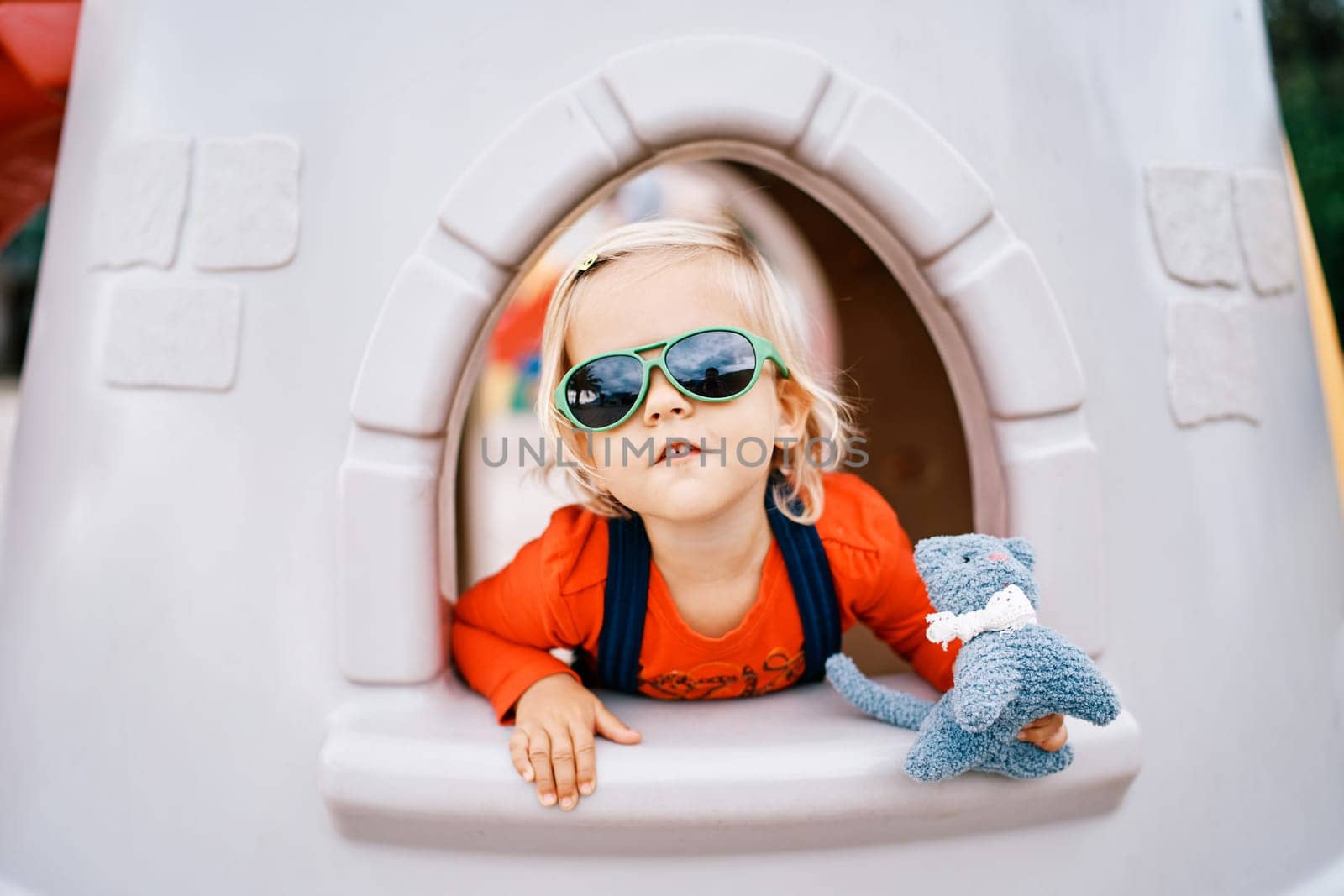Little girl in sunglasses with a toy in her hand looks out of the window of a toy castle on the playground by Nadtochiy