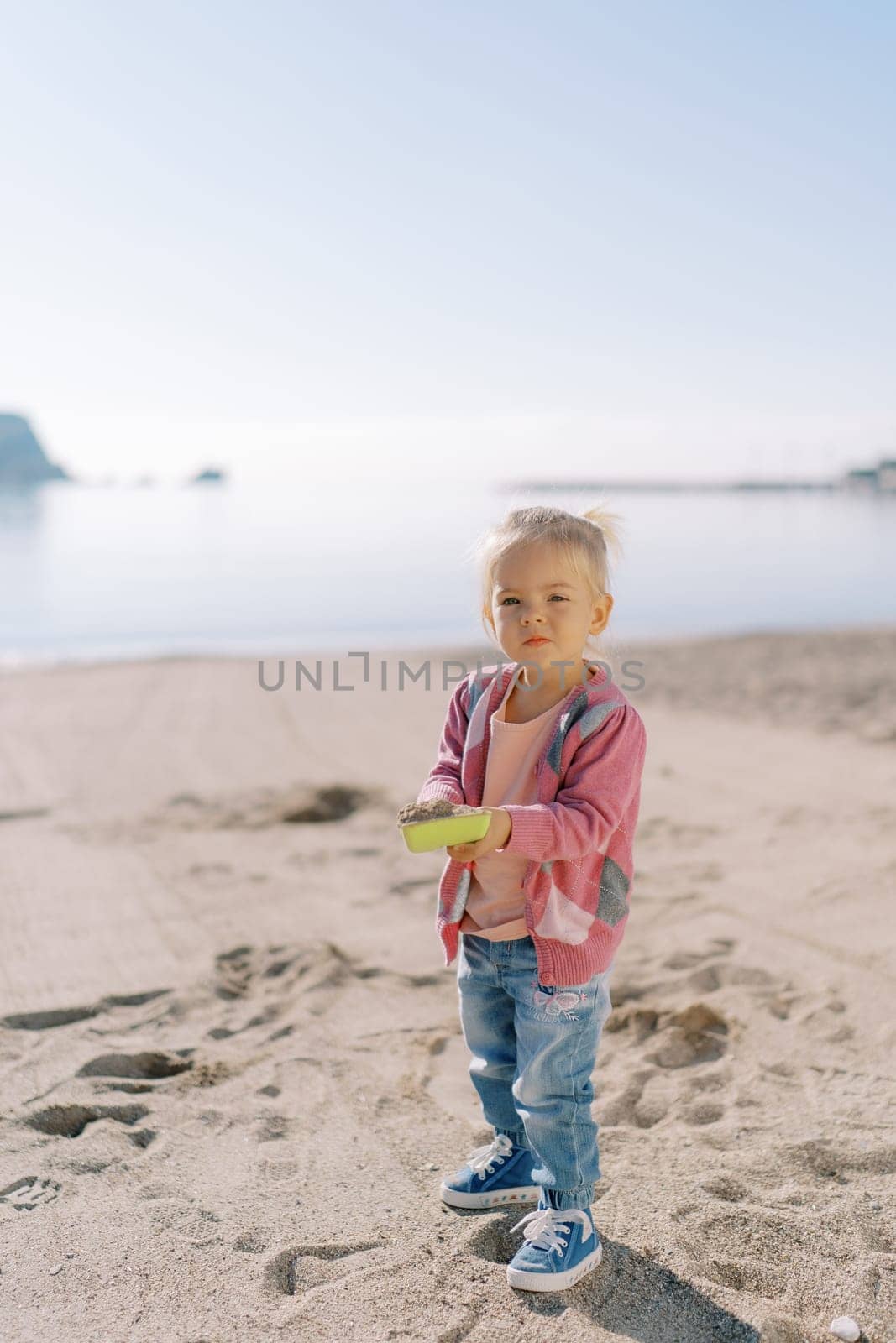 Little girl stands on the beach with a toy shovel full of sand. High quality photo