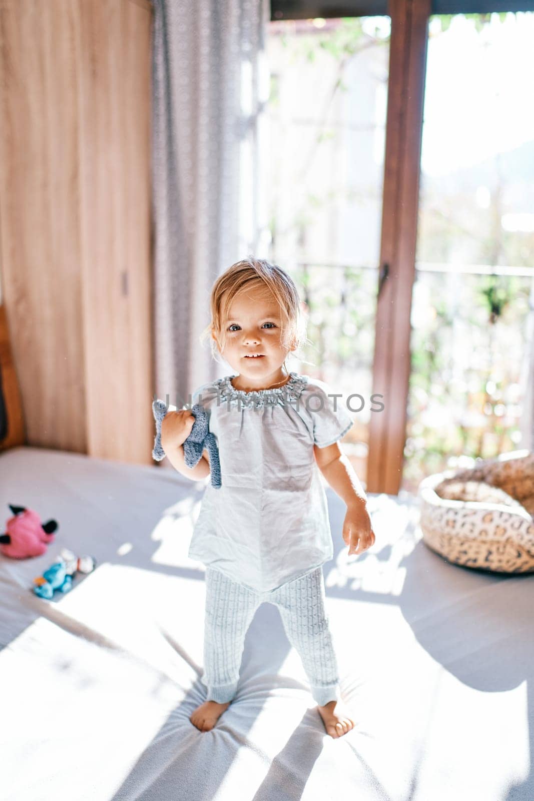 Little smiling girl stands on the bed with a toy under her arm. High quality photo