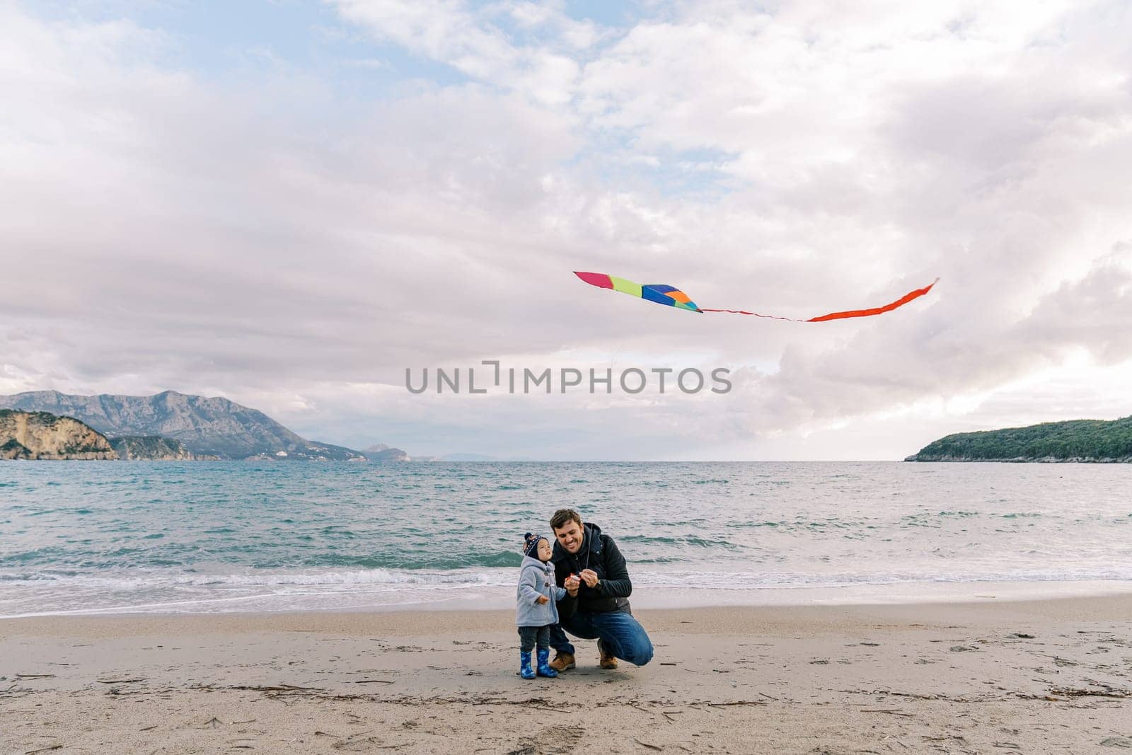Dad, squatting down, launches a colorful kite by the sea with a little girl. High quality photo
