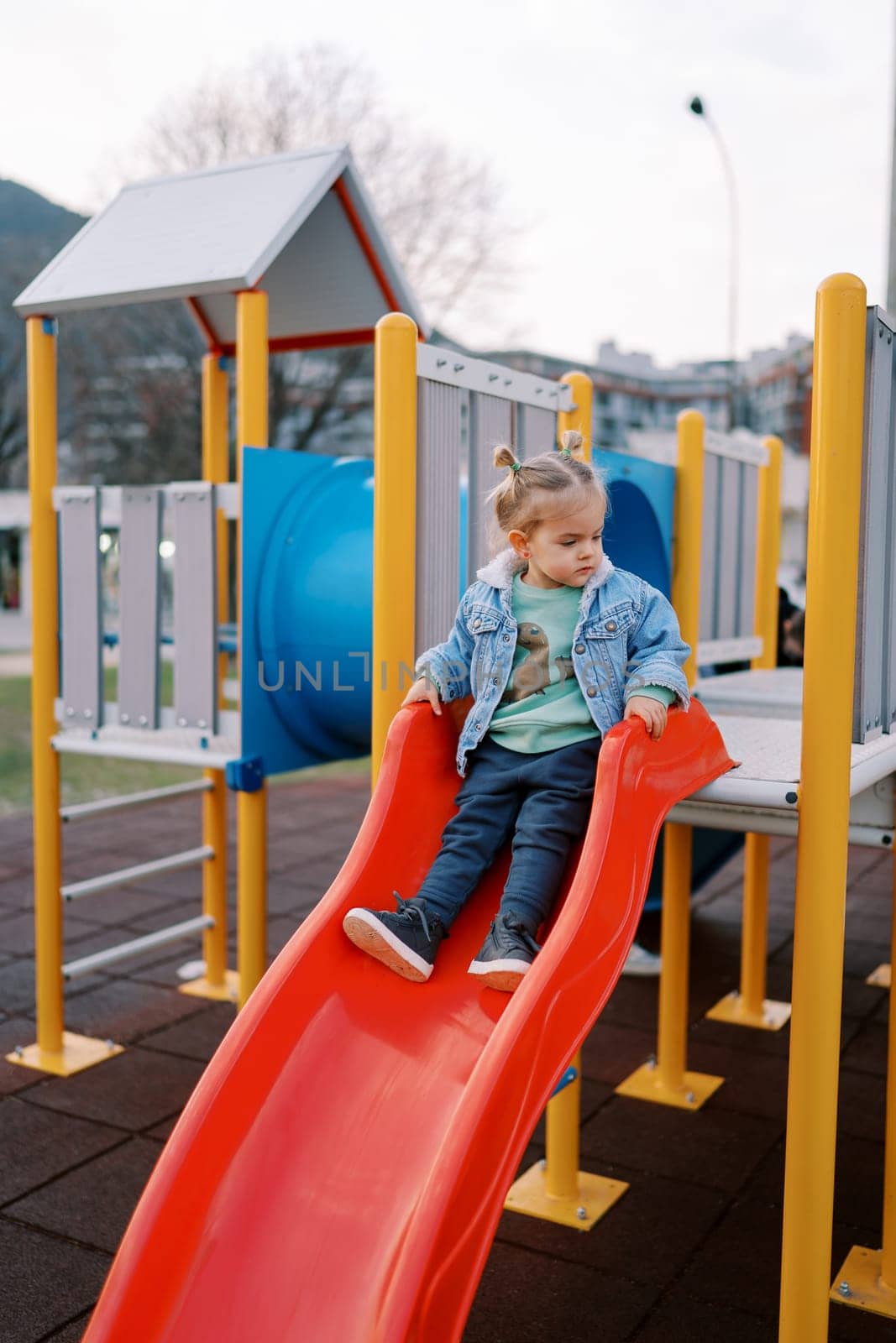 Little girl sits on top of a slide, holding on to the railing and looking away. High quality photo