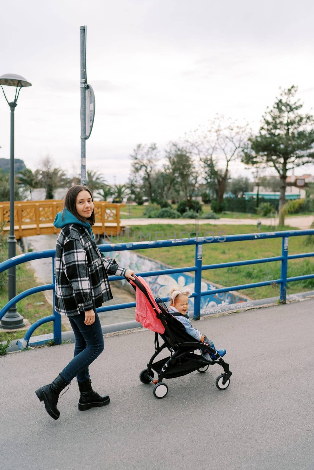 Smiling mother carries a little girl in a stroller along the bridge over the canal in the park. High quality photo