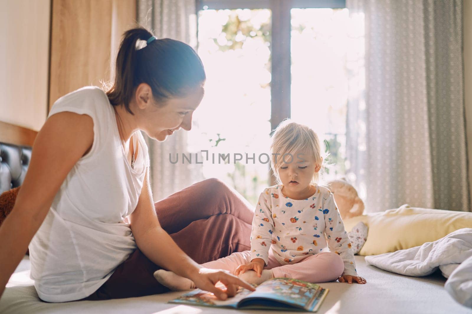 Mom reads a colorful book to a little girl sitting on the bed by Nadtochiy