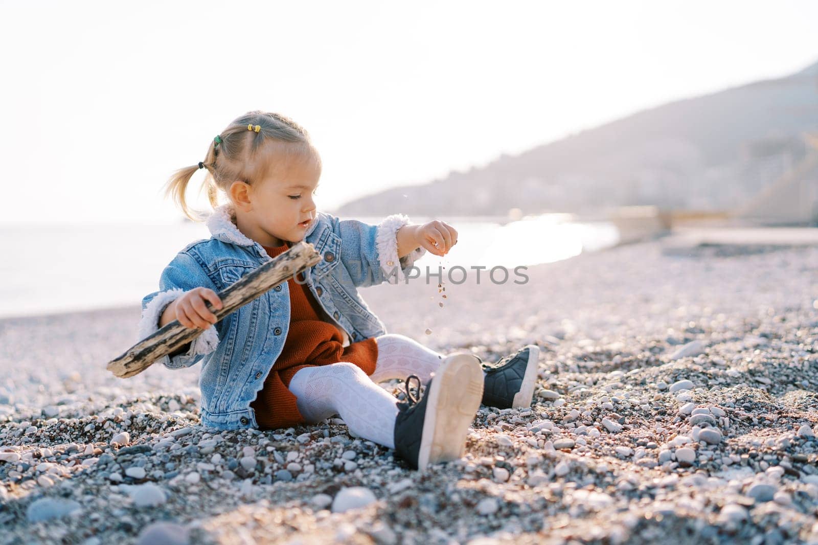 Little girl sits on the beach with a branch and sprinkles small pebbles. Side view. High quality photo