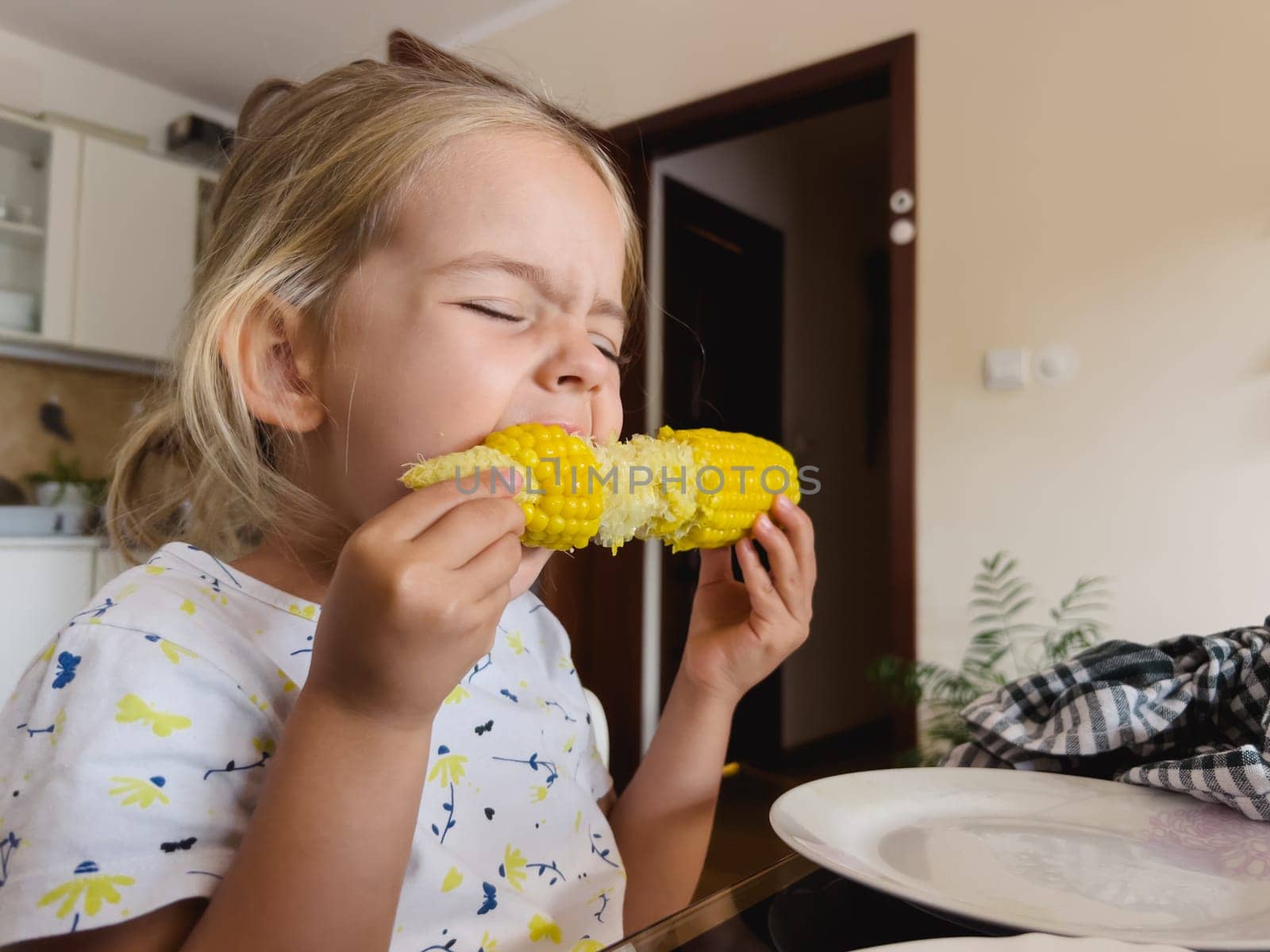 Little girl eating corn on the cob sitting at the table in the kitchen by Nadtochiy