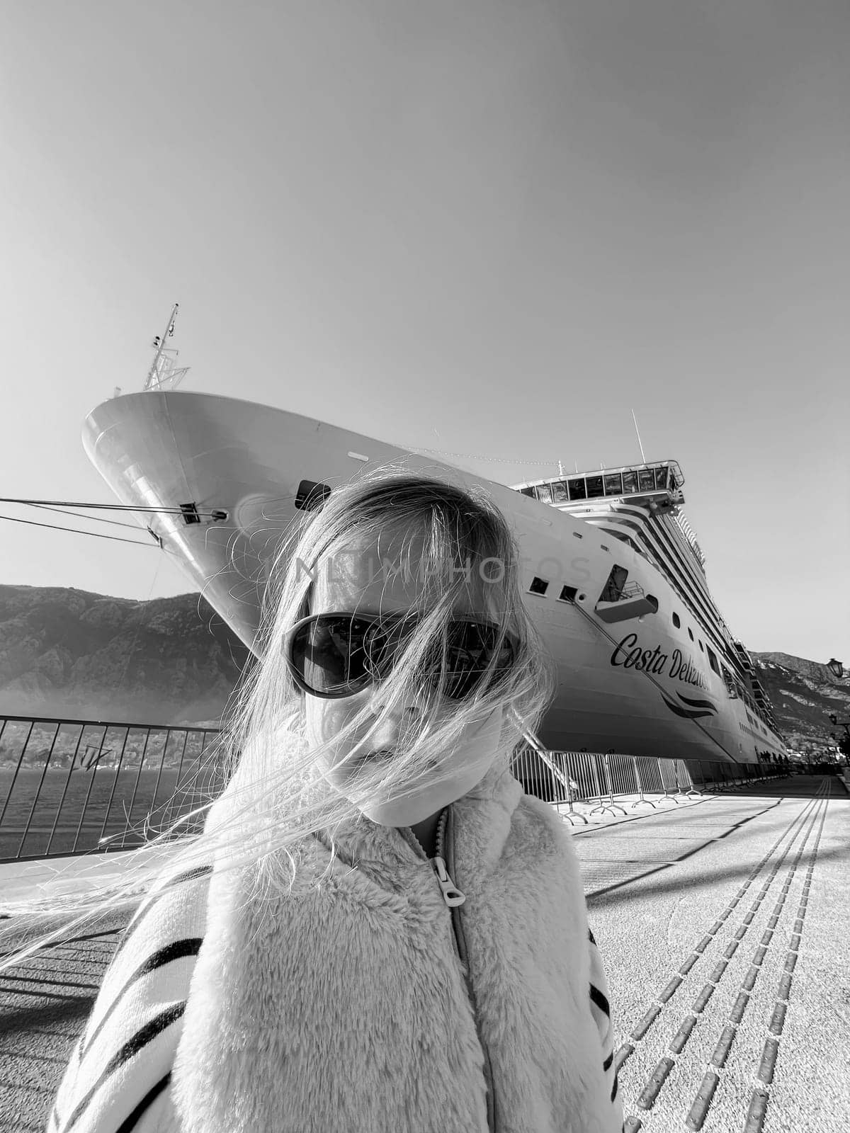 Little girl in sunglasses with flying hair on the background of a cruise liner. Black and white photo by Nadtochiy