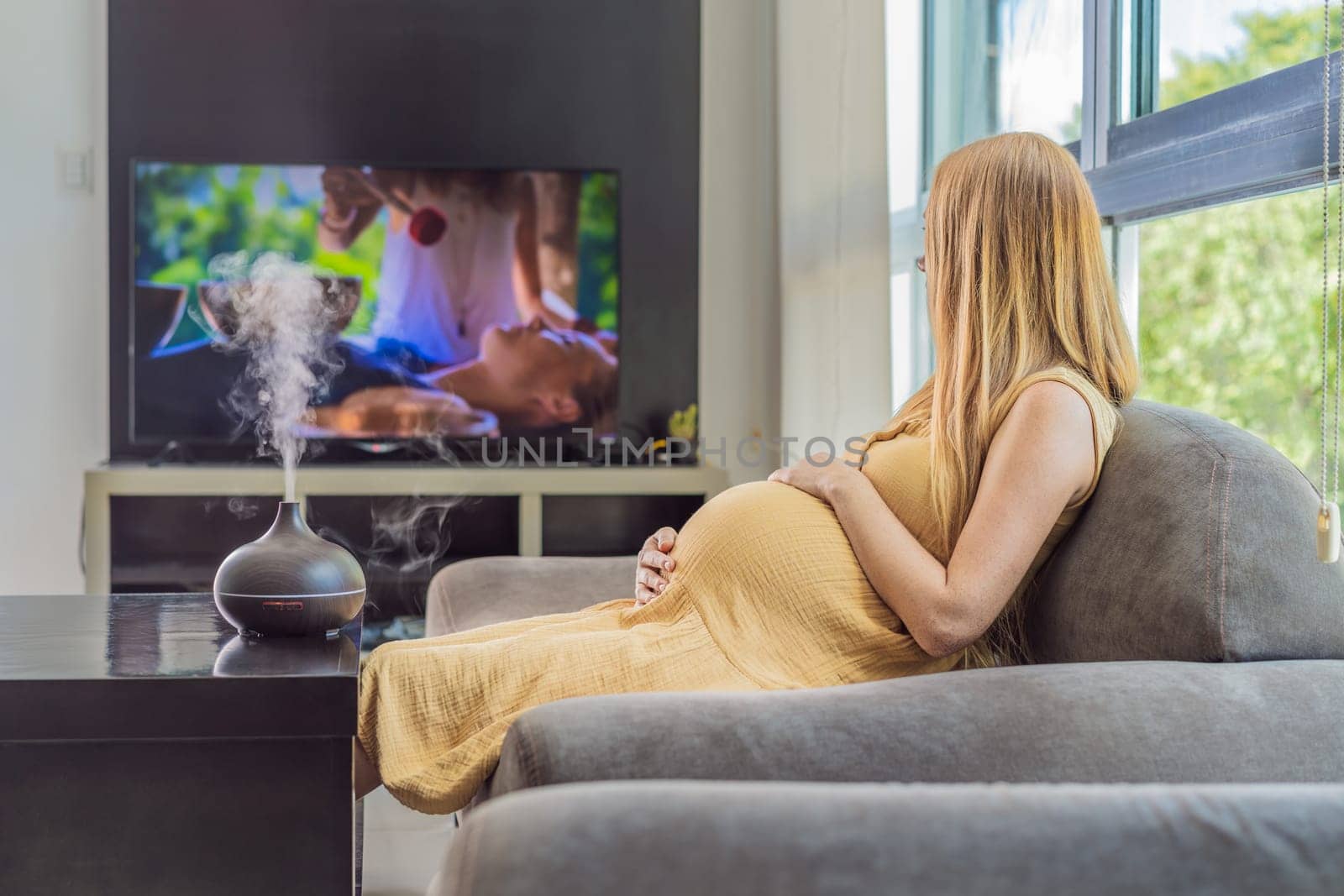 A blissful pregnant woman immerses in relaxation, savoring the soothing aroma from a diffuser while indulging in a calming TV video, embracing tranquility during her pregnant journey by galitskaya