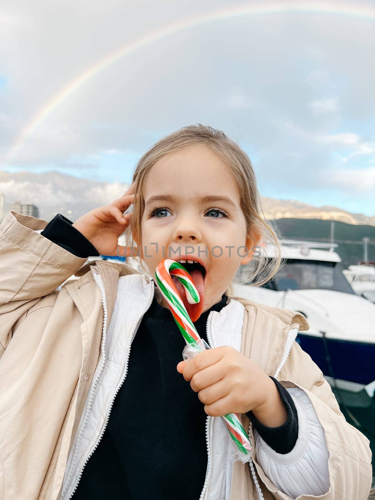 Little girl licks a lollipop against the backdrop of a rainbow over the mountains by Nadtochiy