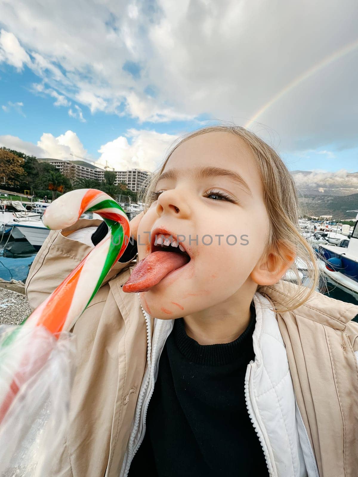 Little girl with her tongue hanging out reaches for a lollipop against the backdrop of a rainbow. High quality photo