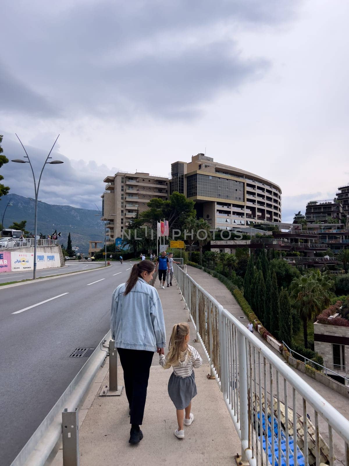 Mom and little girl walk along the sidewalk along the highway to high-rise buildings. Back view. High quality photo