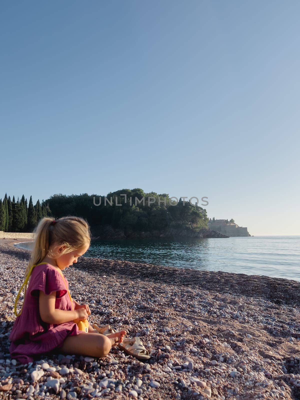 Little girl is sitting on a pebble beach by the sea and opening a small bag. High quality photo