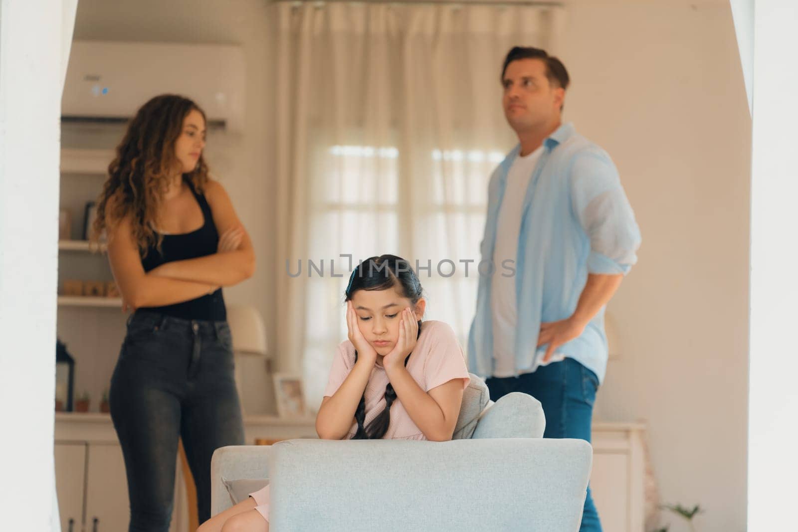 Annoyed and unhappy young girl sitting on sofa trapped in middle of tension by her parent argument in living room. Unhealthy domestic lifestyle and traumatic childhood develop to depression.Synchronos