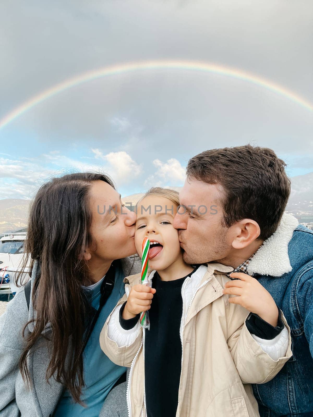 Dad and mom kiss on the cheeks a little girl with a lollipop on a rainbow background by Nadtochiy