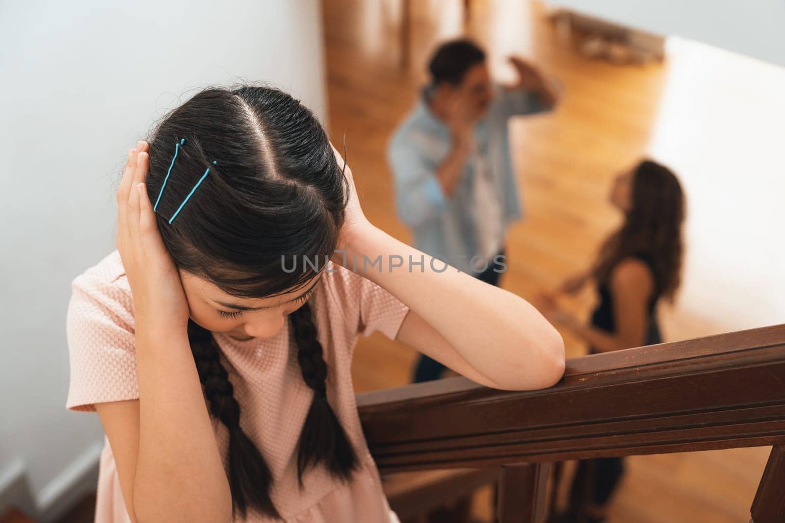 Stressed and unhappy young girl cover her ears blocking sound tension by her parent argument from the stair. Unhealthy family and domestic violence lead to traumatic childhood and anxiety. Synchronos