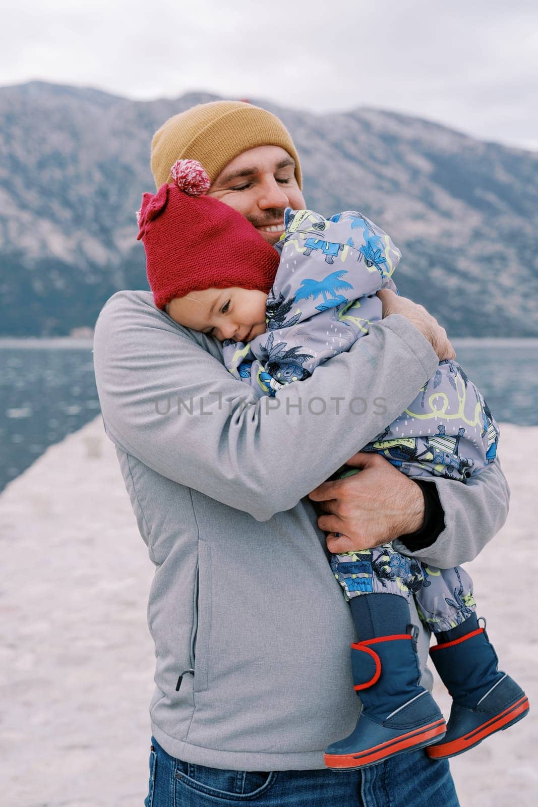 Smiling dad cuddles a little girl on a pier against the backdrop of mountains by Nadtochiy