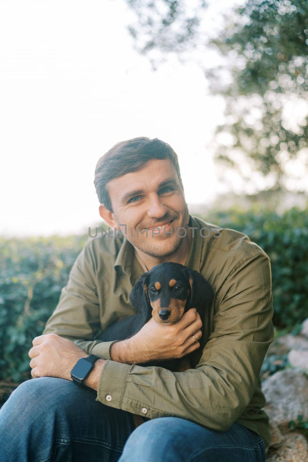 Young smiling man sitting in the park with a black puppy on his lap. High quality photo